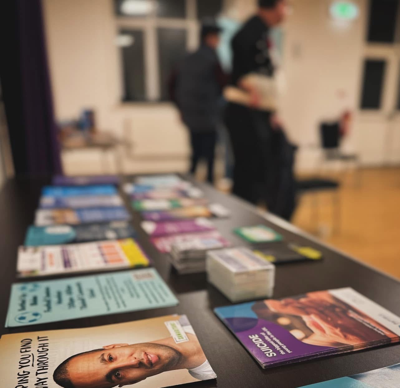 Mental-health information leaflets on a table at a Bottled Up Blokes peer-support group, with attendees intentionally out of focus in the background.